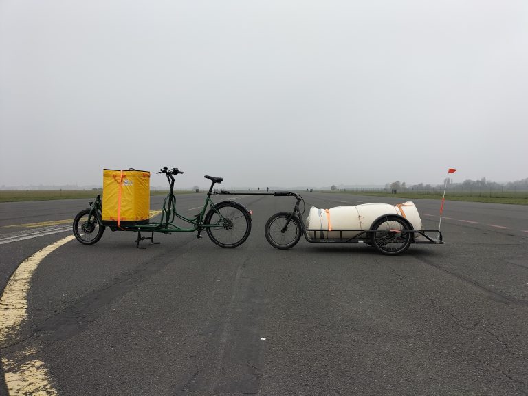 Lastenrad mit großem Fahrradanhänger beladen mit einer zusammengerollten Matratze. Das Rad steht auf einer großen Straße. Der Himmel ist grau.